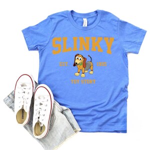 May include: A light blue t-shirt featuring the word "SLINKY" in orange block letters, with a cartoon image of Slinky Dog from Toy Story. The shirt includes the text "EST. 1995" and "TOY STORY". White sneakers and denim shorts are also in the image.