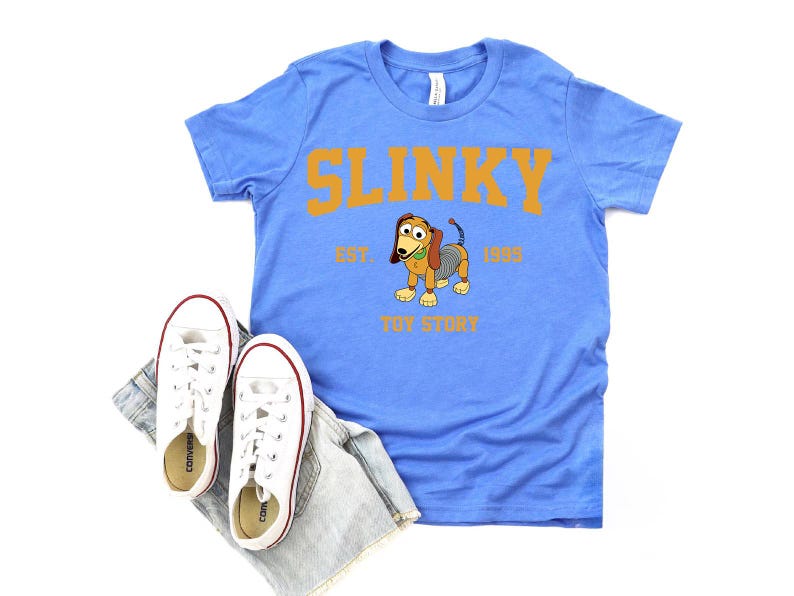 May include: A light blue t-shirt featuring the word "SLINKY" in orange block letters, with a cartoon image of Slinky Dog from Toy Story. The shirt includes the text "EST. 1995" and "TOY STORY". White sneakers and denim shorts are also in the image.