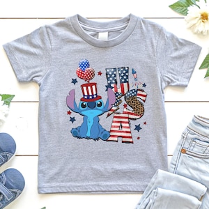K&ouml;nnte beinhalten: Ein hellgraues T-Shirt mit einer blauen Cartoonfigur, die einen patriotischen Hut tr&auml;gt und Luftballons h&auml;lt. Das Shirt hat die Buchstaben "USA" in Rot, Wei&szlig; und Blau, mit einem Leopardenmuster "S". Sterne und ein Feuerwerk vervollst&auml;ndigen das Design.