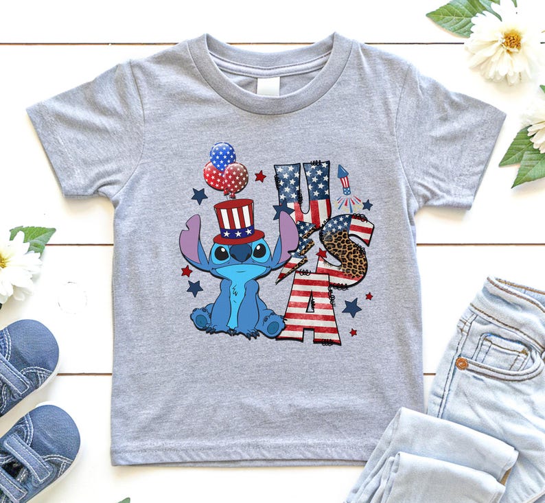K&ouml;nnte beinhalten: Ein hellgraues T-Shirt mit einer blauen Cartoonfigur, die einen patriotischen Hut tr&auml;gt und Luftballons h&auml;lt. Das Shirt hat die Buchstaben "USA" in Rot, Wei&szlig; und Blau, mit einem Leopardenmuster "S". Sterne und ein Feuerwerk vervollst&auml;ndigen das Design.