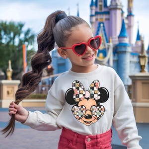 Puede incluir: Una sudadera blanca de manga larga con un gr&aacute;fico de un personaje de rat&oacute;n de dibujos animados que lleva gafas en forma de coraz&oacute;n y un lazo, ambos con un patr&oacute;n de tablero de ajedrez blanco y negro y flores, junto a pantalones rosa y gafas rojas.