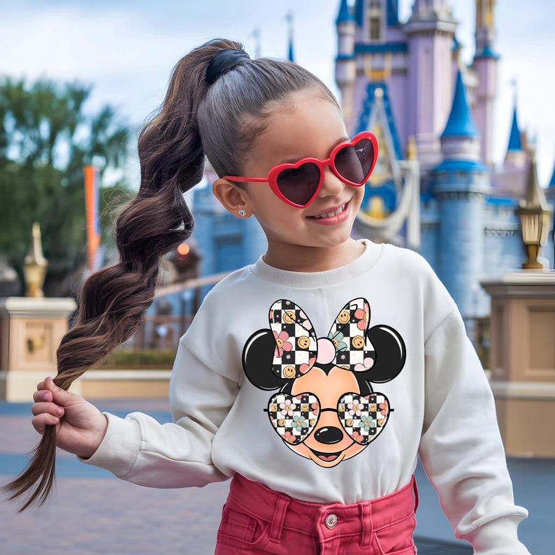 Puede incluir: Una sudadera blanca de manga larga con un gr&aacute;fico de un personaje de rat&oacute;n de dibujos animados que lleva gafas en forma de coraz&oacute;n y un lazo, ambos con un patr&oacute;n de tablero de ajedrez blanco y negro y flores, junto a pantalones rosa y gafas rojas.