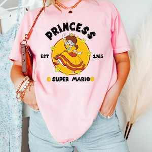 May include: Pink t-shirt featuring a graphic of Princess Daisy from Super Mario. The design includes the word "PRINCESS" above the character, with "EST 1985" and "SUPER MARIO" below. The princess is in a yellow dress.