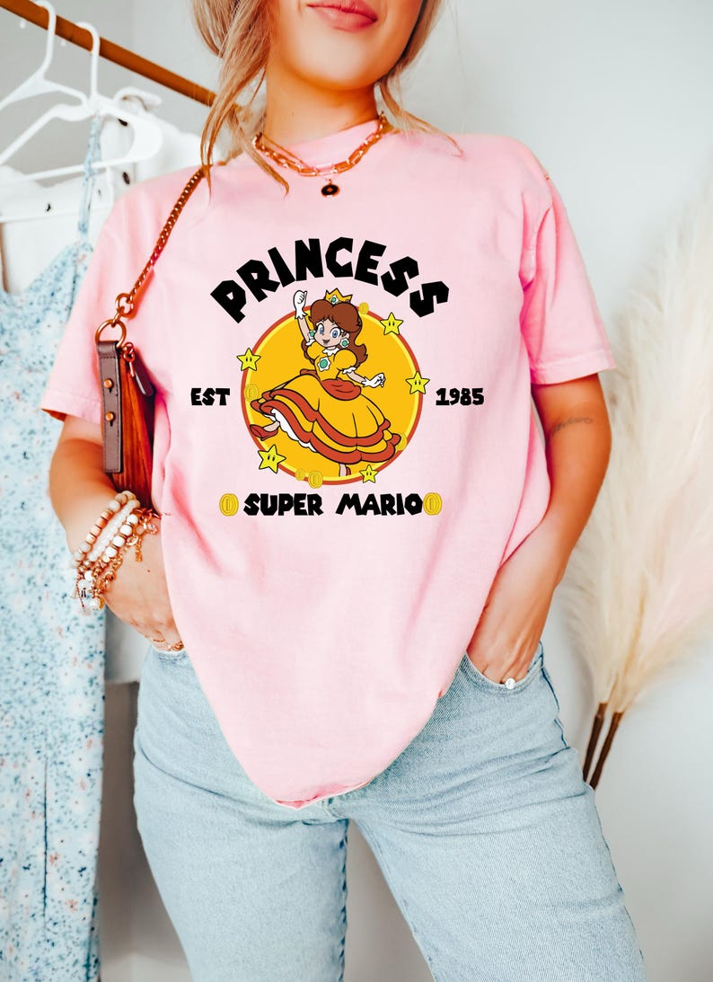 May include: Pink t-shirt featuring a graphic of Princess Daisy from Super Mario. The design includes the word "PRINCESS" above the character, with "EST 1985" and "SUPER MARIO" below. The princess is in a yellow dress.