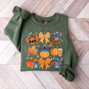 Puede incluir: Una sudadera verde oliva con un dise&ntilde;o tem&aacute;tico de oto&ntilde;o. El dise&ntilde;o incluye personajes de dibujos animados, calabazas, lazos naranjas y hojas de oto&ntilde;o. El gr&aacute;fico est&aacute; centrado en la parte delantera de la sudadera.