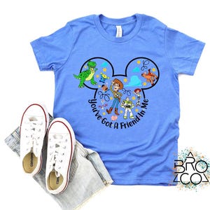 May include: A light blue t-shirt featuring a graphic of Disney's Toy Story characters within a Mickey Mouse ear design. The text "You've Got a Friend in Me" is also included. White sneakers and denim shorts are also in the image.