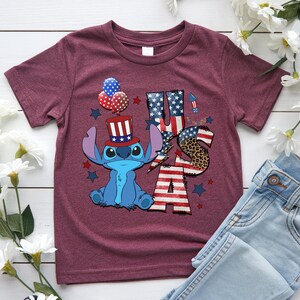 K&ouml;nnte beinhalten: Burgunderfarbenes T-Shirt mit einer blauen Cartoonfigur, die einen patriotischen Hut tr&auml;gt, und den Buchstaben "USA" in Rot, Wei&szlig; und Blau. Das Design umfasst Sterne, Luftballons und ein Leopardenmuster.