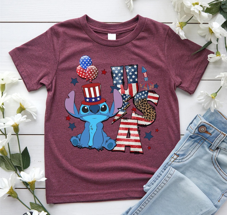 K&ouml;nnte beinhalten: Burgunderfarbenes T-Shirt mit einer blauen Cartoonfigur, die einen patriotischen Hut tr&auml;gt, und den Buchstaben "USA" in Rot, Wei&szlig; und Blau. Das Design umfasst Sterne, Luftballons und ein Leopardenmuster.