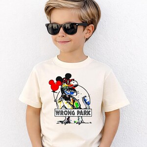 May include: A cream-colored short-sleeve t-shirt featuring a graphic of a colorful T-Rex wearing red sunglasses and Mickey Mouse ears. The dinosaur holds a red Mickey-shaped balloon and stands behind a sign that reads WRONG PARK in black text.