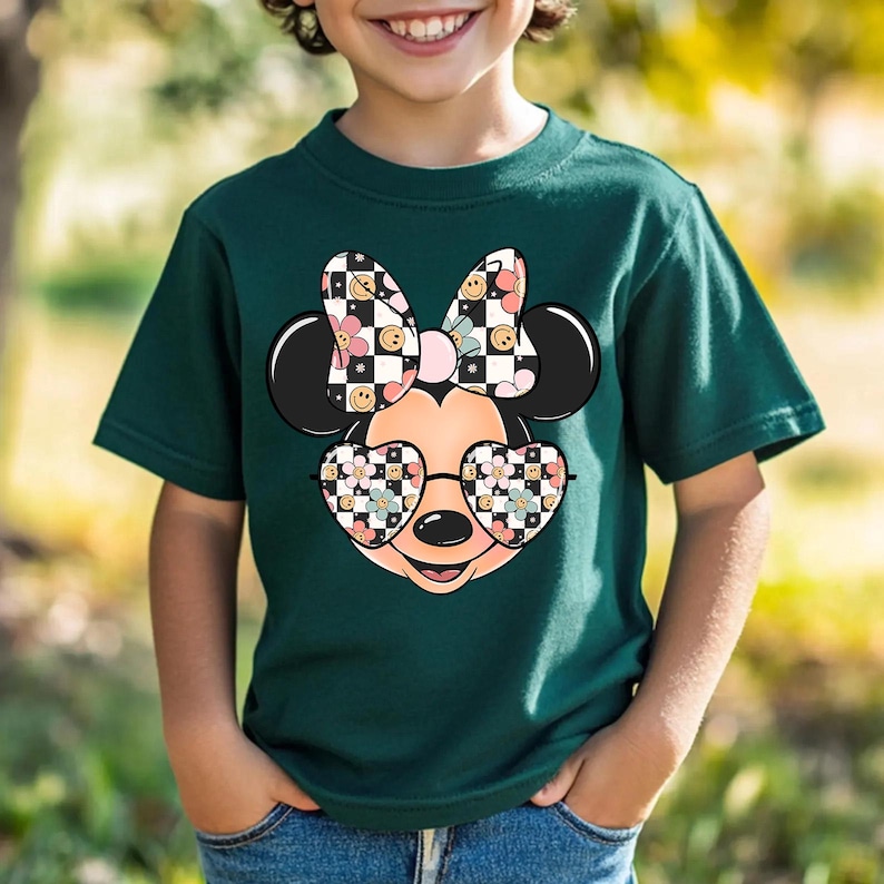Puede incluir: Una camiseta de manga corta de color verde oscuro con un gr&aacute;fico de una cabeza de rat&oacute;n de dibujos animados con gafas en forma de coraz&oacute;n y un lazo. El lazo y las gafas tienen un patr&oacute;n de tablero de ajedrez, caritas sonrientes y flores.