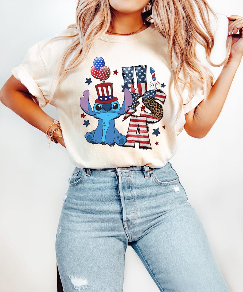 K&ouml;nnte beinhalten: Cremefarbenes T-Shirt mit einer blauen Cartoonfigur, die einen patriotischen Hut tr&auml;gt, und dem Text "USA" in Rot, Wei&szlig; und Blau. Das Design umfasst Sterne, Feuerwerk und ein Leopardenmuster. Das Shirt wird mit hellblauen Jeans getragen.