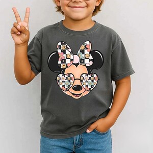 Puede incluir: Una camiseta gris carb&oacute;n de manga corta con un gr&aacute;fico de una cabeza de rat&oacute;n de dibujos animados que lleva gafas en forma de coraz&oacute;n y un lazo. El lazo y las gafas tienen un patr&oacute;n de tablero de ajedrez blanco y negro, flores rosas y caritas felices amarillas.