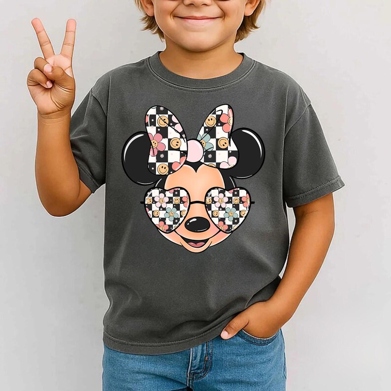 Puede incluir: Una camiseta gris carb&oacute;n de manga corta con un gr&aacute;fico de una cabeza de rat&oacute;n de dibujos animados que lleva gafas en forma de coraz&oacute;n y un lazo. El lazo y las gafas tienen un patr&oacute;n de tablero de ajedrez blanco y negro, flores rosas y caritas felices amarillas.