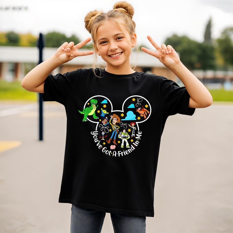 May include: Black t-shirt featuring a graphic of Disney's Toy Story characters inside a Mickey Mouse head outline. The text "You've Got A Friend In Me" is written in a circular pattern around the characters. The girl is wearing gray jeans.