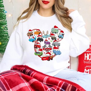 May include: White sweatshirt featuring a Cars-themed design with various cartoon cars wearing Santa hats. The design is arranged in a Mickey Mouse shape, with the word "Cars" and a checkered flag. A Christmas tree and holiday decor are in the background.