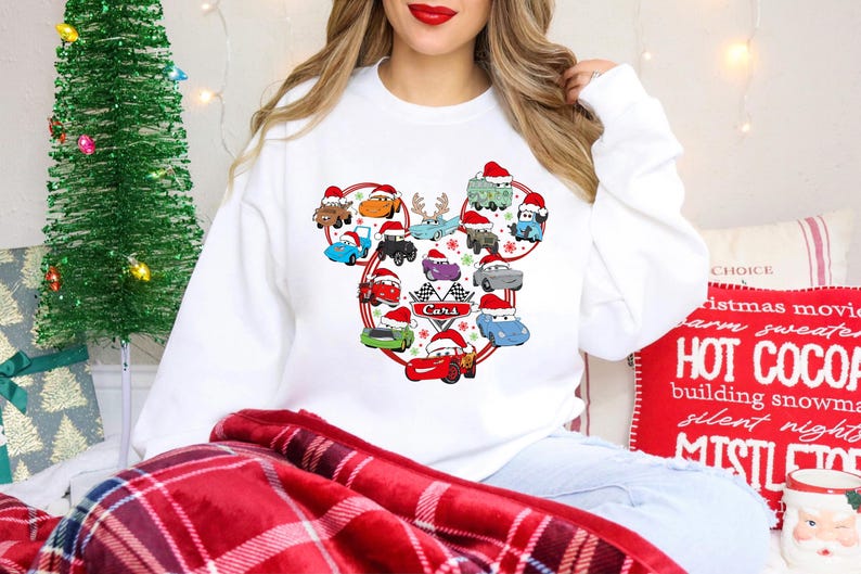 May include: White sweatshirt featuring a Cars-themed design with various cartoon cars wearing Santa hats. The design is arranged in a Mickey Mouse shape, with the word "Cars" and a checkered flag. A Christmas tree and holiday decor are in the background.