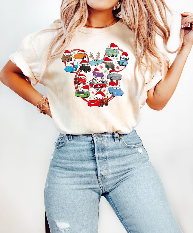 May include: Cream-colored t-shirt featuring a graphic of various Cars characters wearing Santa hats, arranged in a Mickey Mouse head shape. The word "Cars" is printed in the center. The shirt is paired with light blue jeans.