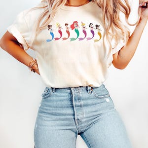 May include: Cream-colored t-shirt featuring a row of cartoon mermaids with colorful tails and hair. The mermaids are in various poses, with different skin tones and hair colors. The shirt is paired with light blue denim jeans.