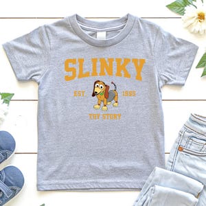 May include: A light gray t-shirt featuring the word "SLINKY" in orange block letters above a cartoon image of Slinky Dog from Toy Story. The text "EST. 1995" and "TOY STORY" are also printed on the shirt.