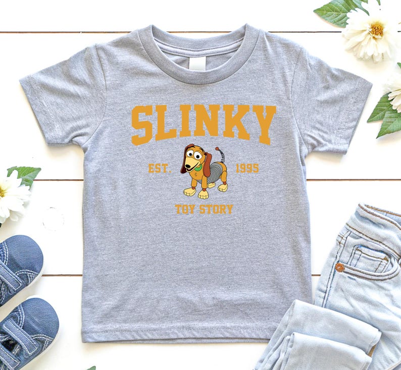 May include: A light gray t-shirt featuring the word "SLINKY" in orange block letters above a cartoon image of Slinky Dog from Toy Story. The text "EST. 1995" and "TOY STORY" are also printed on the shirt.