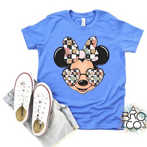 Puede incluir: Una camiseta de manga corta azul claro con un gr&aacute;fico de una cabeza de rat&oacute;n de dibujos animados que lleva gafas en forma de coraz&oacute;n y un lazo. El lazo y las gafas est&aacute;n decorados con un patr&oacute;n de tablero de ajedrez, flores y caras sonrientes.