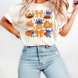 Puede incluir: Camiseta color crema con un dise&ntilde;o tem&aacute;tico de Halloween. El dise&ntilde;o incluye personajes de dibujos animados, calabazas, hojas de oto&ntilde;o y lazos naranjas. La camiseta se combina con vaqueros azul claro.