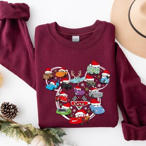 May include: Burgundy sweatshirt featuring a Cars-themed design. The graphic showcases various cartoon cars wearing Santa hats, arranged in a Mickey Mouse-like shape. The word "Cars" is printed in white. The sweatshirt has a crew neck and long sleeves.