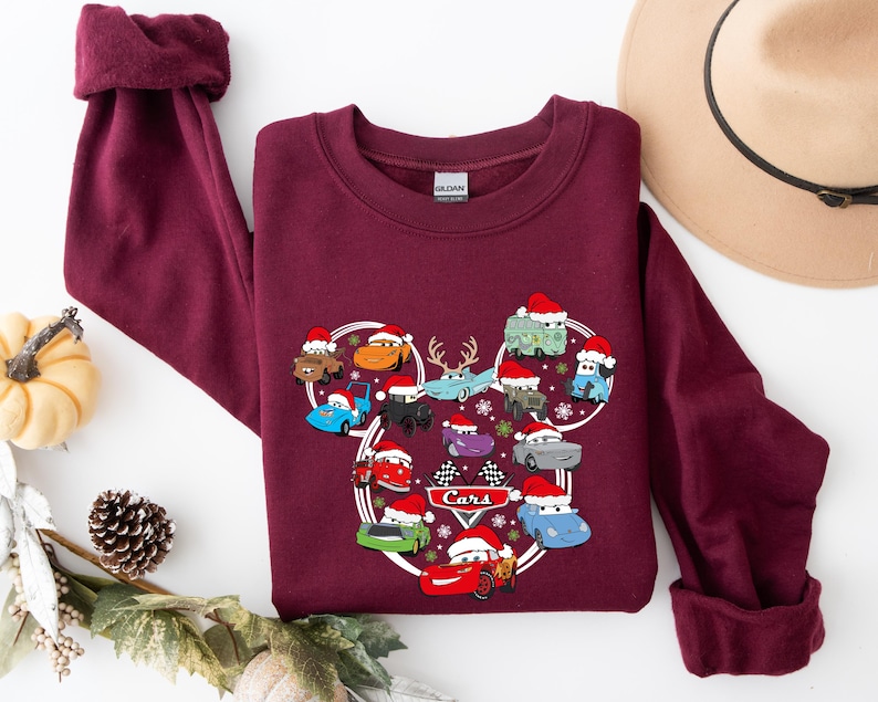 May include: Burgundy sweatshirt featuring a Cars-themed design. The graphic showcases various cartoon cars wearing Santa hats, arranged in a Mickey Mouse-like shape. The word "Cars" is printed in white. The sweatshirt has a crew neck and long sleeves.