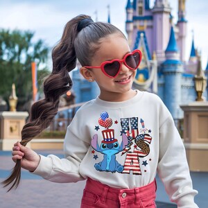 K&ouml;nnte beinhalten: Ein cremefarbenes Sweatshirt mit einem patriotischen Stitch-Design und dem Text "USA". Das Design umfasst Sterne, Streifen und eine Zeichentrickfigur mit Hut. Die Person tr&auml;gt eine rote herzf&ouml;rmige Sonnenbrille und eine rote Hose.