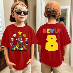 K&ouml;nnte beinhalten: Rotes T-Shirt mit einem Super Mario-Motiv und dem Text "Super Birthday" auf der Vorderseite. Auf der R&uuml;ckseite stehen der Name "KEVIN" und die Zahl "8" in Gelb. Das T-Shirt wird von einem Kind getragen.