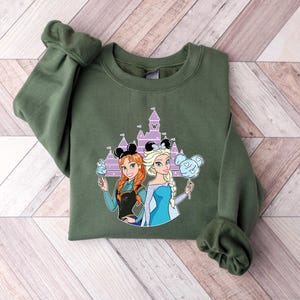 May include: An olive green sweatshirt featuring a graphic of two cartoon princesses with a castle in the background. The princesses are holding treats and wearing mouse ears. The sweatshirt is made of a soft material.