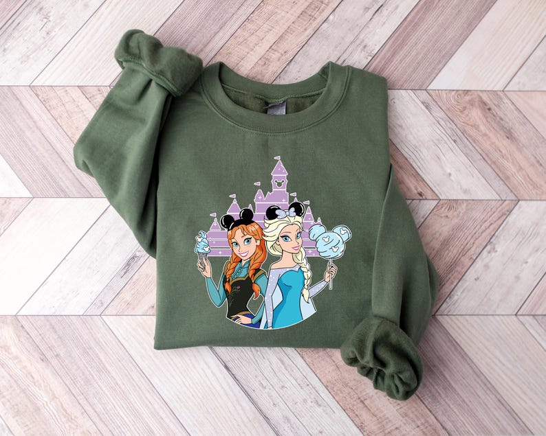 May include: An olive green sweatshirt featuring a graphic of two cartoon princesses with a castle in the background. The princesses are holding treats and wearing mouse ears. The sweatshirt is made of a soft material.