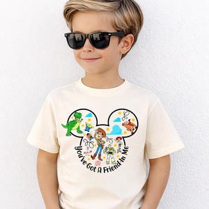 May include: A cream-colored t-shirt featuring a graphic of Disney's Toy Story characters within a Mickey Mouse head outline. The text "You've Got A Friend In Me" is printed below the characters. The shirt is worn by a child wearing black sunglasses.