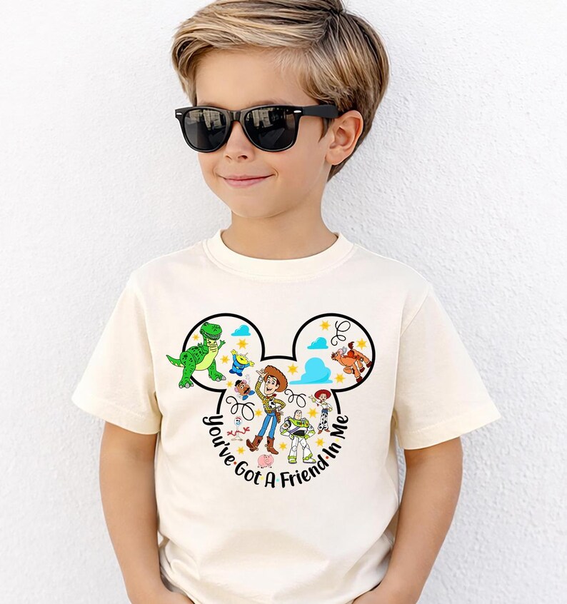 May include: A cream-colored t-shirt featuring a graphic of Disney's Toy Story characters within a Mickey Mouse head outline. The text "You've Got A Friend In Me" is printed below the characters. The shirt is worn by a child wearing black sunglasses.
