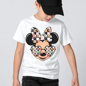 Puede incluir: Una camiseta blanca de manga corta con un gr&aacute;fico de una cabeza de rat&oacute;n de dibujos animados que lleva gafas en forma de coraz&oacute;n y un lazo grande. El lazo y las gafas est&aacute;n decorados con un patr&oacute;n de tablero de ajedrez blanco y negro, flores rosas y caras sonrientes amarillas.