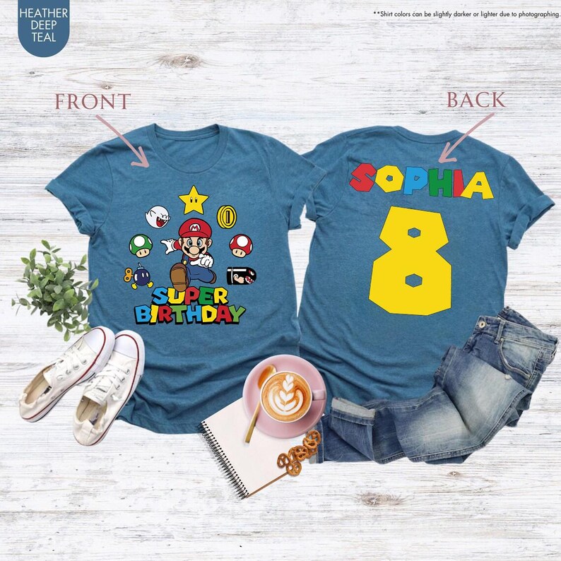 K&ouml;nnte beinhalten: T&uuml;rkisfarbene T-Shirts mit einem Super Mario-Motiv. Das vordere Shirt zeigt Mario und die Worte "Super Birthday". Das hintere Shirt hat den Namen "Sophia" und die Zahl "8" in Gelb. Die Shirts werden mit Jeans und Schuhen pr&auml;sentiert.