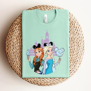 May include: A mint green t-shirt featuring cartoon illustrations of two princesses with mouse ear headbands. One holds an ice cream cone, the other a cotton candy. A purple castle silhouette is in the background.