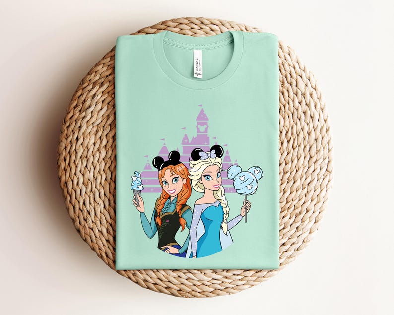 May include: A mint green t-shirt featuring cartoon illustrations of two princesses with mouse ear headbands. One holds an ice cream cone, the other a cotton candy. A purple castle silhouette is in the background.