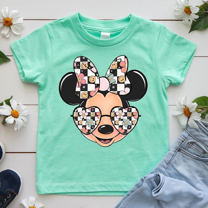 Puede incluir: Una camiseta de manga corta color verde menta con el gr&aacute;fico de una cabeza de rat&oacute;n de dibujos animados que lleva gafas de sol en forma de coraz&oacute;n. El lazo y las gafas tienen un patr&oacute;n de tablero de ajedrez blanco y negro, caritas sonrientes amarillas y flores rosas y azules.