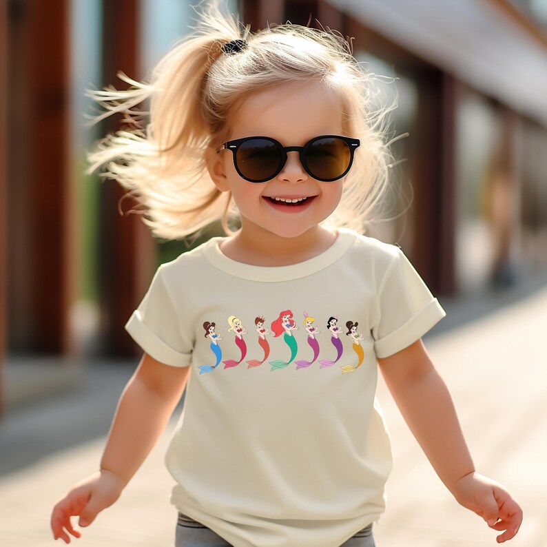 May include: A young child wearing a cream-colored t-shirt with a colorful mermaid graphic. The graphic features several mermaids with different hair colors and tail colors. The child is also wearing black sunglasses.