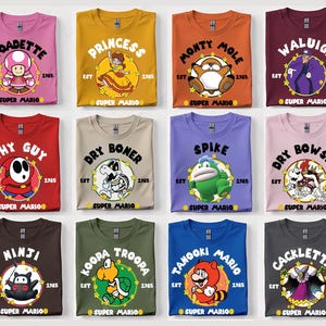 May include: A collection of Super Mario themed t-shirts in various colors, each featuring a different character graphic and the text "EST 1985 SUPER MARIO". Characters include Toadette, Princess, Monty Mole, Waluigi, Shy Guy, Dry Boner, Spike, Dry Bowser, Ninji, Koopa Troopa, Tanooki Mario, and Cackletta.