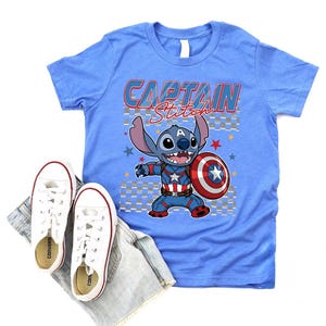 May include: A light blue t-shirt with the words "CAPTAIN Stitch" and a cartoon character holding a shield. White sneakers and denim shorts are placed below the shirt. The shirt features a graphic of Stitch as Captain America.