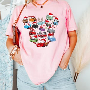 May include: Pink t-shirt featuring a graphic of cartoon cars wearing Santa hats, arranged in a Mickey Mouse head shape. The word "Cars" is printed below a checkered flag. The shirt is a light pink color.