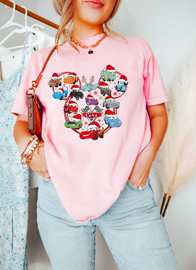May include: Pink t-shirt featuring a graphic of cartoon cars wearing Santa hats, arranged in a Mickey Mouse head shape. The word "Cars" is printed below a checkered flag. The shirt is a light pink color.