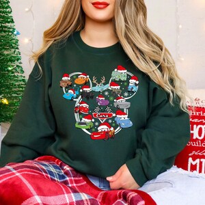 May include: Dark green sweatshirt featuring a graphic of cartoon cars wearing Santa hats, arranged in a Mickey Mouse head shape. The word "Cars" is in the center. A Christmas tree is in the background.