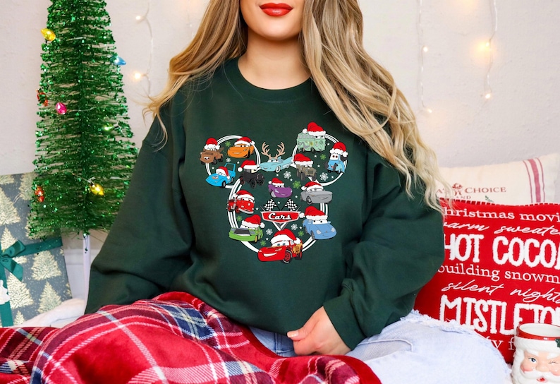 May include: Dark green sweatshirt featuring a graphic of cartoon cars wearing Santa hats, arranged in a Mickey Mouse head shape. The word "Cars" is in the center. A Christmas tree is in the background.