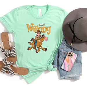 May include: A light green t-shirt featuring a cartoon image of Sheriff Woody riding a horse, with the text "Sheriff Woody". The shirt is paired with denim shorts, a gray hat, sandals, and a phone case.