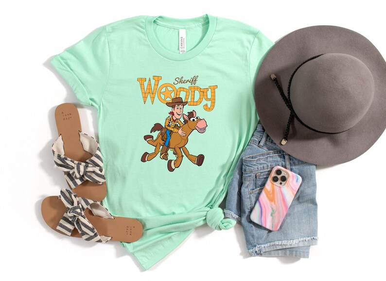 May include: A light green t-shirt featuring a cartoon image of Sheriff Woody riding a horse, with the text "Sheriff Woody". The shirt is paired with denim shorts, a gray hat, sandals, and a phone case.