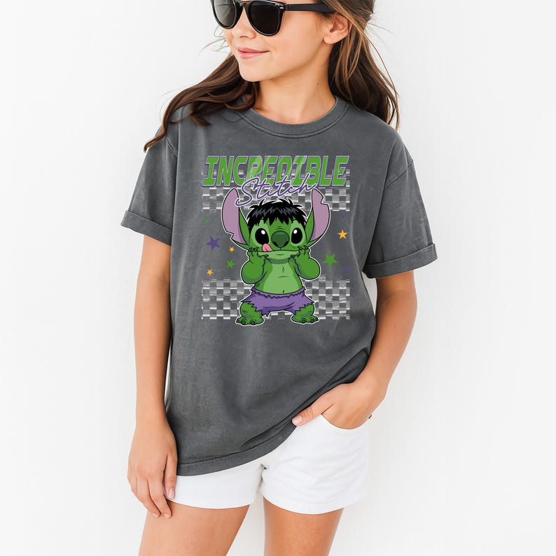May include: Dark gray t-shirt featuring a graphic of a green character with purple ears and shorts, styled as a mashup of Stitch and the Hulk. The text "Incredible Stitch" is above the character in green and purple.