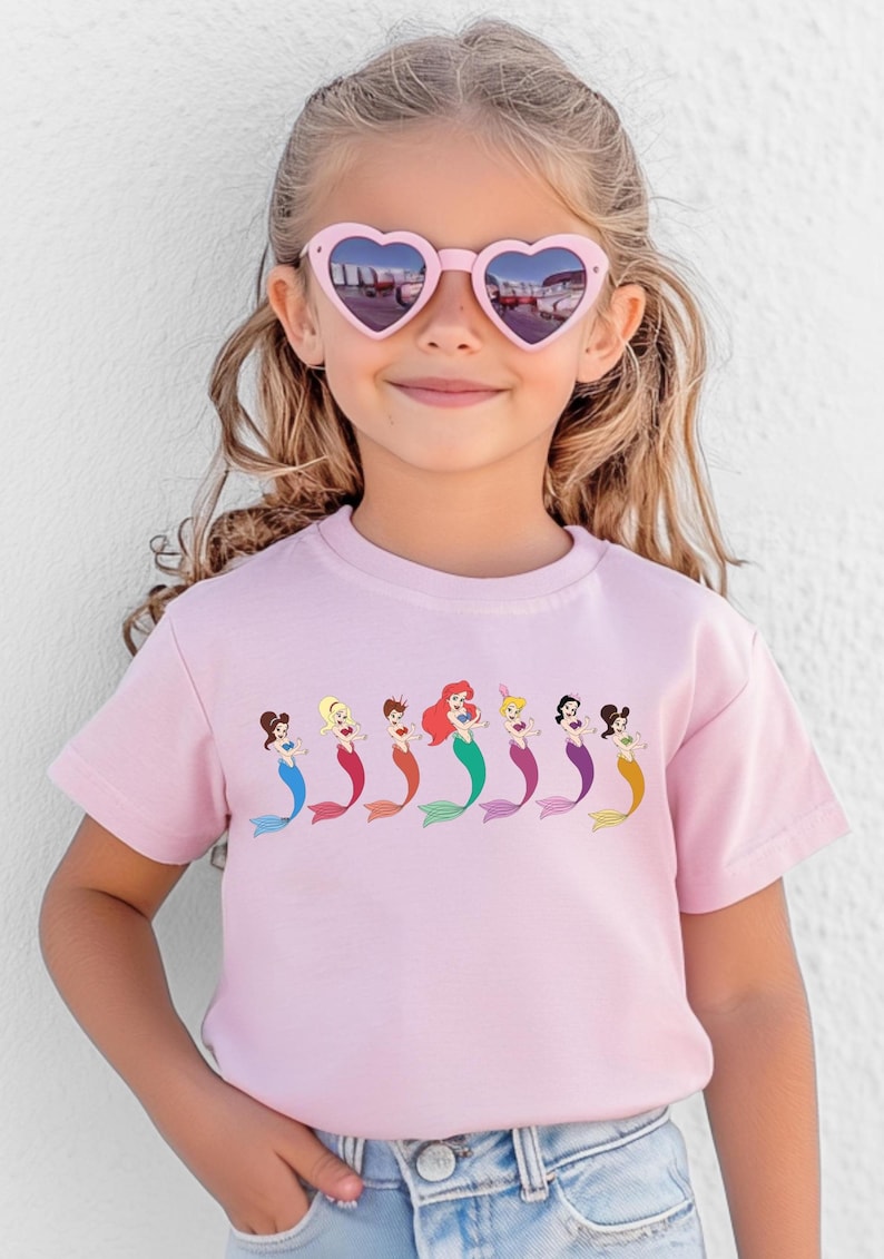 May include: A light pink t-shirt featuring a row of cartoon mermaids with colorful tails. The mermaids have different hair colors and styles. The girl in the image is wearing pink heart-shaped sunglasses.